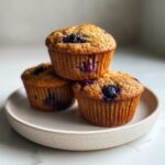 Three perfectly baked blueberry oatmeal muffins stacked on a speckled ceramic plate.