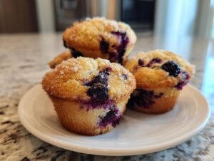 Three delicious blueberry oatmeal muffins piled on a white plate, featuring a coarse sugar topping and visible blueberries.
