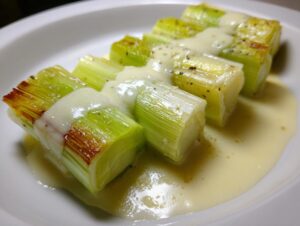 Four segments of tender braised leeks topped with a creamy white sauce, served on a white plate.