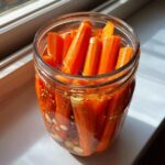 Close-up of bright orange pickled carrots packed tightly in a glass jar with brine, garlic, and mustard seeds.