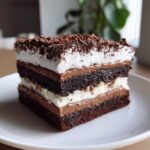 A decadent slice of brownie refrigerator cake featuring multiple layers of dark cake, chocolate mousse, and white cream, topped with chocolate shavings.