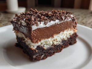 A close-up of a decadent slice of brownie refrigerator cake showing a dark brownie base, white layer, chocolate mousse, and chocolate shavings.