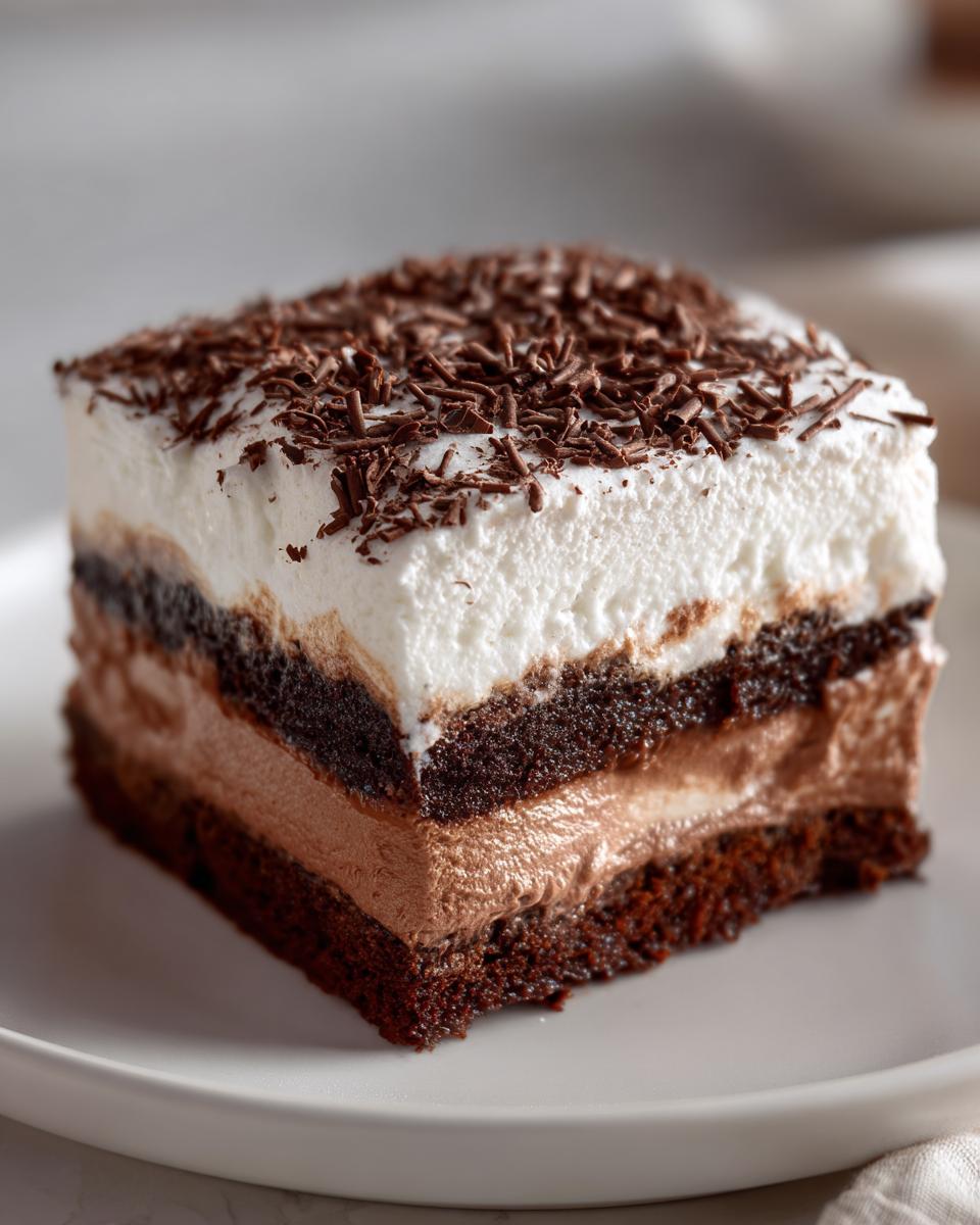 A square slice of rich brownie refrigerator cake showing layers of dark cake, chocolate mousse, and white whipped cream topped with chocolate shavings.