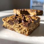 A close-up, sunlit view of a thick, chewy chocolate chip oatmeal bars square on a white counter.