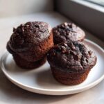 Three dark, moist chocolate zucchini muffins topped with chocolate chips sitting on a white plate.