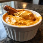 A pretzel rod is dipped into a small white ramekin filled with creamy, orange beer cheese pub dip.