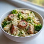 Close-up of creamy chicken sausage and broccoli orzo served in a white bowl, featuring browned sausage slices and bright green florets.