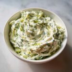 A white bowl filled with creamy dill pickle dip, featuring visible chunks of pickle and fresh dill mixed throughout.