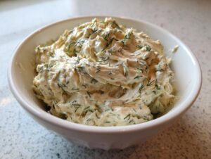 A close-up of creamy, textured dill pickle dip generously garnished with fresh dill in a white bowl.