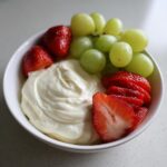 A white bowl filled with creamy fruit dip, surrounded by fresh green grapes and sliced strawberries.