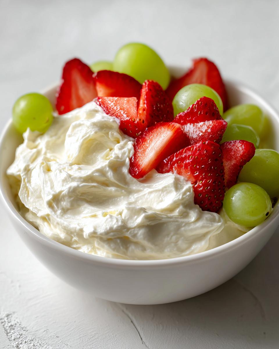 A white bowl filled with creamy fruit dip, topped with fresh sliced strawberries and green grapes.