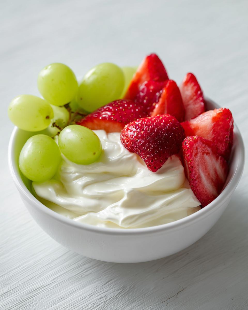 Close-up of a white bowl filled with creamy fruit dip, topped with fresh strawberries and green grapes.