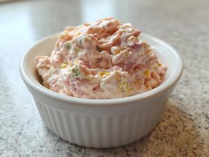 A close-up of a creamy portion of ham salad recipe served in a small, white, fluted ramekin on a speckled countertop.