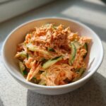 A white bowl filled with vibrant orange and white kani salad mixed with thinly sliced cucumbers and topped with sesame seeds.