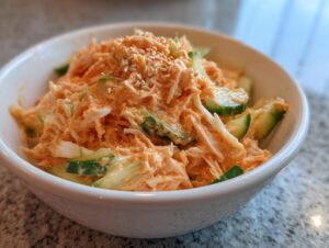 Close-up of creamy, orange-tinted kani salad mixed with sliced cucumbers and topped with sesame seeds.