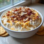 A white bowl filled with creamy Neiman Marcus dip, topped generously with crumbled bacon and sliced almonds.