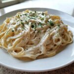 A close-up of fettuccine pasta generously coated in creamy Olive Garden Alfredo sauce, topped with black pepper and parsley.