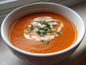 A close-up of a white bowl filled with rich, orange tomato bisque, swirled with cream and topped with fresh basil.