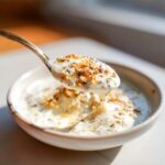A spoonful of thick, creamy white pizza sauce topped with everything bagel seasoning being lifted from a small bowl.