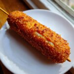 A perfectly fried, golden brown korean corn dog on a stick resting on a white plate.