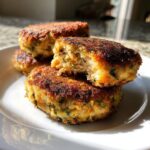 Close-up of golden-brown, crispy salmon cakes stacked on a white plate, one cut open showing flaky texture.