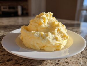 A generous mound of creamy, yellow mashed potatoes served on a white plate, highlighted by sunlight.