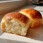 Close-up of two golden-brown japanese milk bread rolls, one sliced open revealing a soft, fluffy interior.