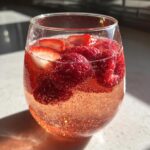 Close-up of a glass filled with bubbly, pink champagne punch garnished with fresh raspberries and sliced strawberries.