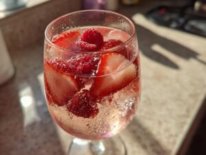 Close-up of a glass filled with bubbly champagne punch, ice, sliced strawberries, and raspberries.