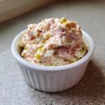 A close-up of a small white ramekin filled high with creamy ham salad recipe mixture, showing chunks of pink ham and green relish.