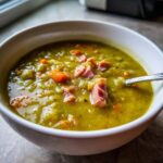 Close-up of a white bowl filled with thick, green pea soup topped with chunks of ham and diced carrots.