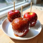 Three shiny, deep red caramel apples on sticks, resting on a white plate.