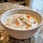 A close-up of a white bowl filled with creamy homemade cream of chicken soup, topped with shredded chicken, carrots, and herbs.