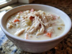 A close-up view of a white bowl filled with rich homemade cream of chicken soup, featuring shredded chicken and diced carrots.