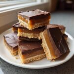 A stack of homemade, layered twix bars featuring shortbread, caramel, and chocolate topping, served on a white plate.