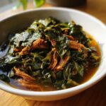 A close-up of tender collard greens simmered in broth with shredded smoked meat served in a white bowl.