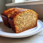 Close-up of a thick slice of moist banana bread recipe loaf with a golden-brown crust, served on a white plate.