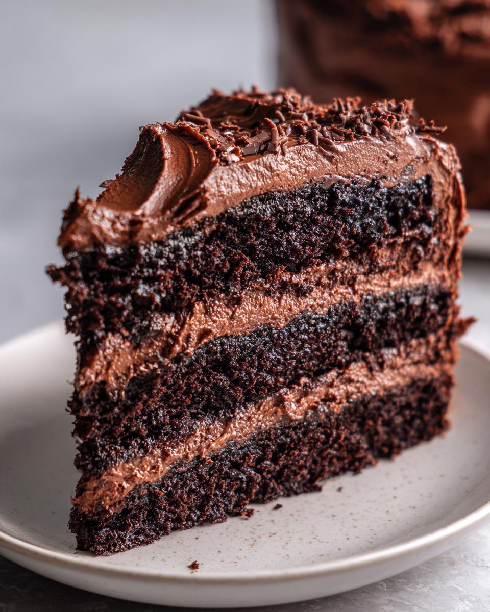 Close-up of a rich, moist slice of chocolate cake with three layers and thick chocolate frosting, topped with chocolate shavings.