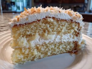 Close-up of a moist slice of coconut cake recipe, layered with white frosting and topped with toasted coconut flakes.