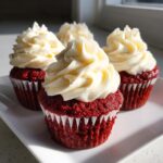 Four perfectly frosted red velvet cupcakes with bright white cream cheese frosting sitting on a white plate.