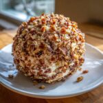 A close-up of a perfectly formed cheese ball recipe coated entirely in chopped pecans and seasonings, resting on a white plate.