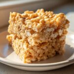 Two stacked squares of golden, chewy peanut butter rice krispie treats sitting on a small white plate.