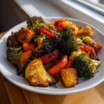 A close-up of perfectly cooked roasted vegetables, including charred broccoli, cauliflower, and red peppers, served on a white plate.