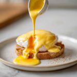 A rich, yellow hollandaise sauce is being poured from a small white pitcher over a poached egg on toast.