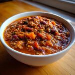 A close-up of a white bowl filled with rich, thick pumpkin chili, showing chunks of orange pumpkin, kidney beans, and ground meat.