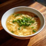 A white bowl filled with hot egg drop soup, featuring delicate egg ribbons and topped generously with fresh chopped green onions.