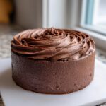 A small round cake completely covered in rich, dark chocolate frosting with piped swirls on top.