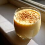A glass of rich, creamy yellow eggnog topped generously with ground cinnamon, catching sunlight.