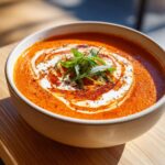 A close-up of a bowl of vibrant orange roasted tomato soup, swirled with cream and topped with fresh green herbs.