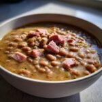 Close-up of a white bowl filled with savory 15 bean soup, topped with chunks of ham.